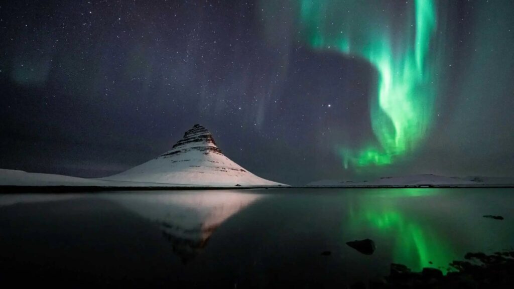 Kirkjufell mountain illuminated by the northern lights, reflecting in a calm lake under a starry night sky.
