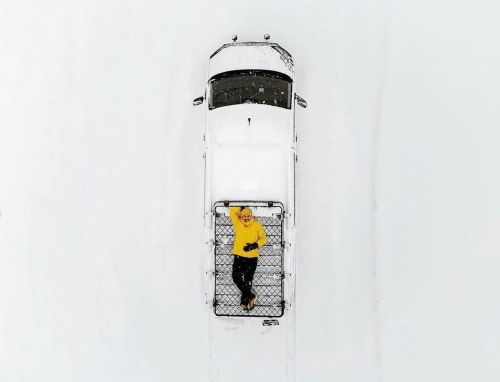 A person in a yellow jacket laying roof rack of a white campervan, surrounded by a white snowy background.