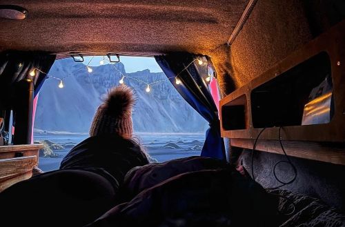 View from the inside of the campervan. A woman is lying on the bedding and watching the Icelandic landscape via opened back of the campervan.