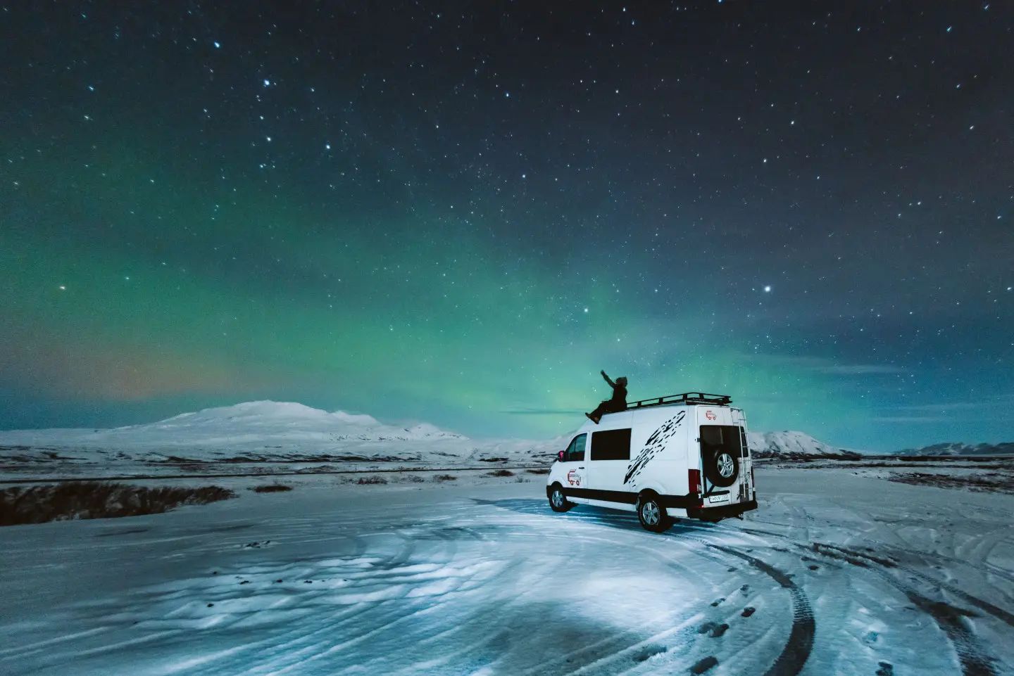 A man is sitting on the roof of a campervan while watching dancing northern lights.