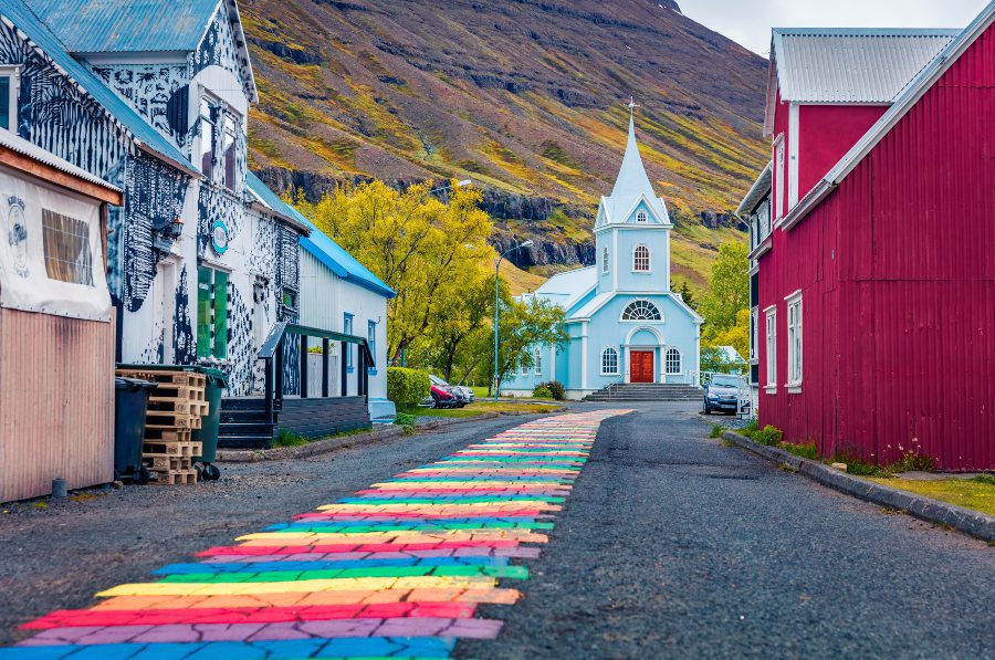 A rainbow road leading to a small blue church in the town of Seyðisfjörður.