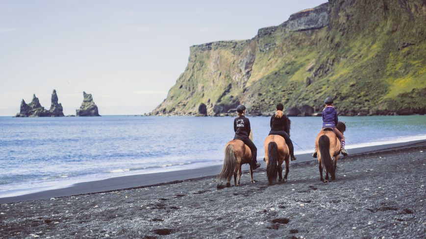 3 people riding a horses on black beach with the sea and the mountains in the background