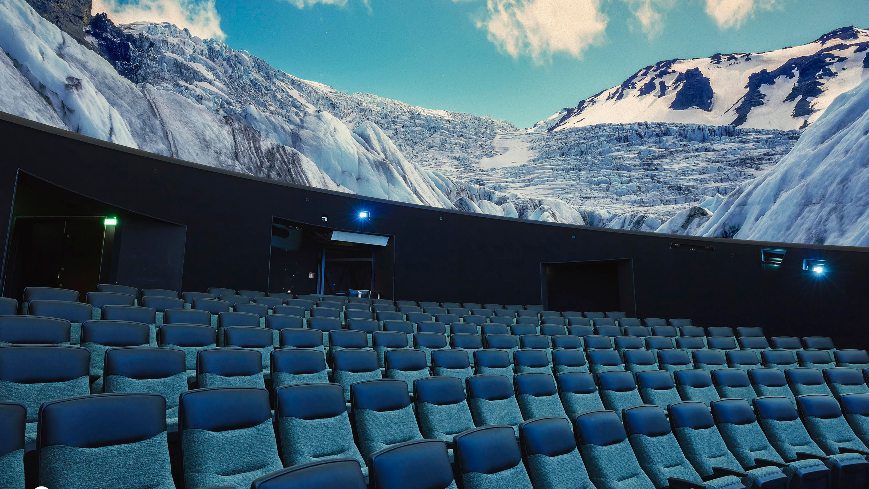Empty hall with many seats. At the top there is screen with view of mountains
