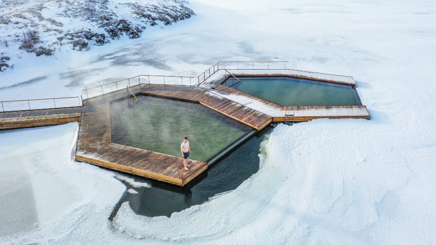 Man standing near geothermal pools next to ice-bound lake
