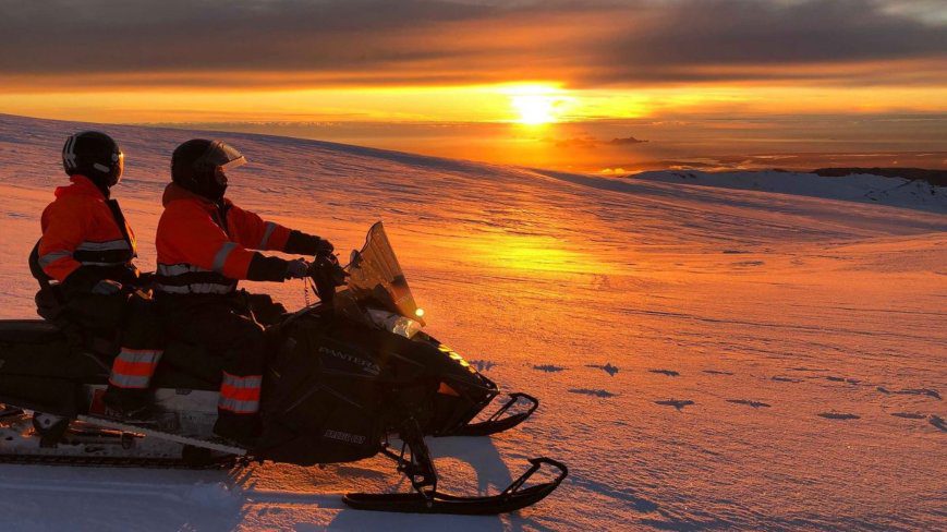 2 people are watching the sunset during their snowmobiling tour. There is a lot of snow