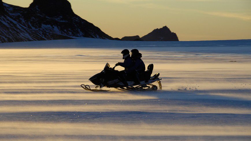 2 people is riding snowmobile on snow. There are the mountains in the background