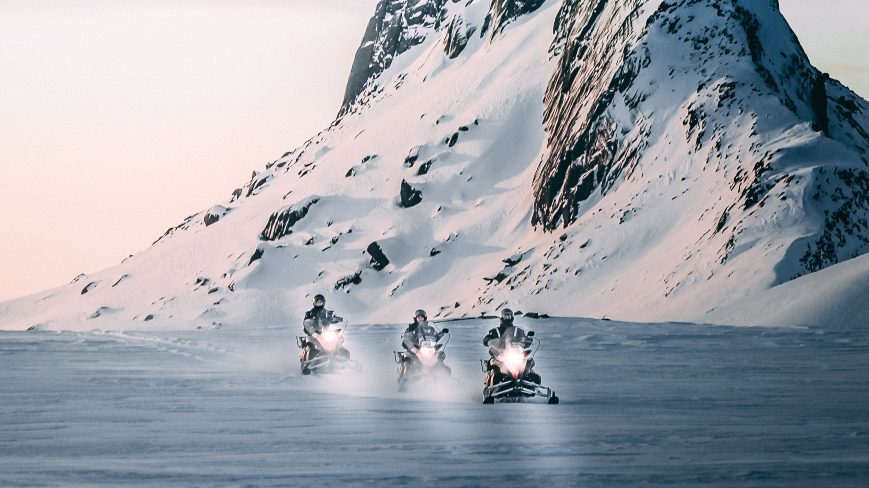 3 snowmobiles is riding through snowy road. There is a mountain in the background