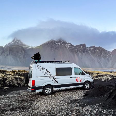Man is standing at the top of a camper van. In the background there are mountains