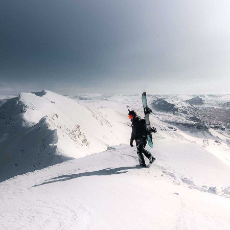 Person is hiking on the snow carrying a skies on his back. In the background there are mountains fully covered by snow