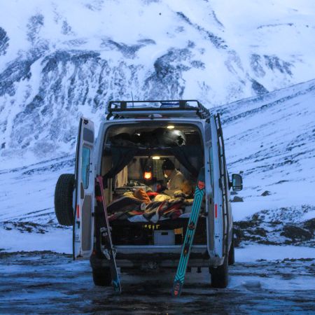A woman is laying inside the camper van with opened doors. In the background there are mountains