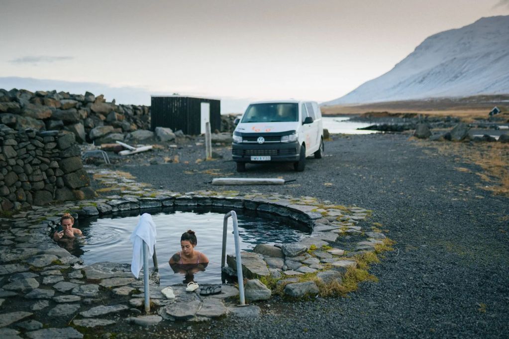 Two individuals relax in a natural hot spring surrounded by rocky terrain, with a parked van nearby and mountains in the background.