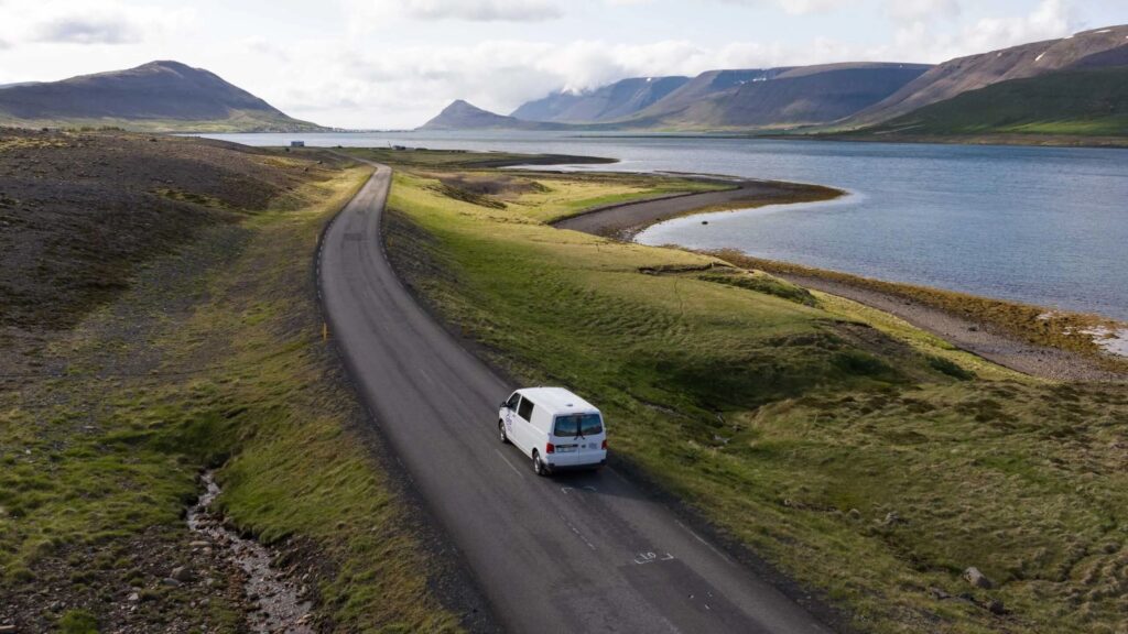A white campervan drives on a scenic road beside a tranquil bay, surrounded by lush green hills and majestic mountains in Iceland.