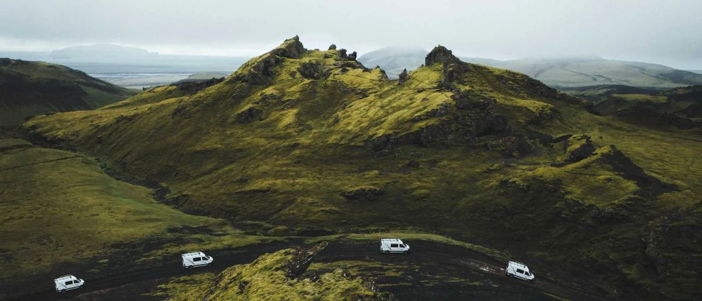 Aerial view of three white vans navigating a winding road through lush green hills and rocky terrain under a cloudy sky.