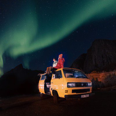 Woman is sitting on the roof of the campervan and watching the Northern Lights