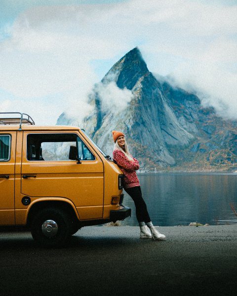 Woman is standing in front of the campervan next to the lake and a big mountain