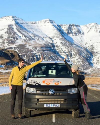 Two people happily posing with their hands on a parked van in front of a snow-capped mountain range.