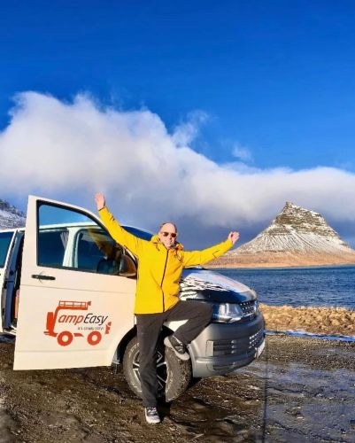 Man in a yellow jacket posing joyfully beside a CampEasy camper van, parked by a scenic lake with a snowy mountain in the background.