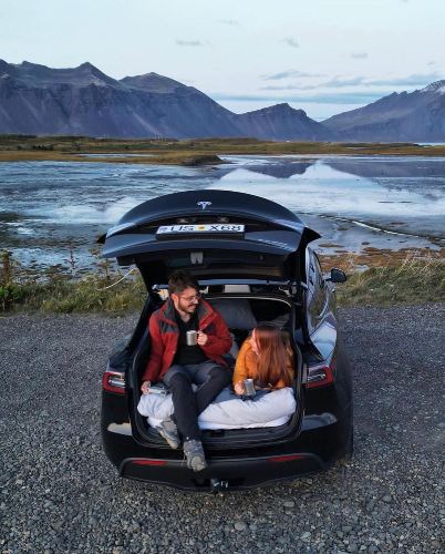Two people sitting in the trunk of a Tesla Model Y, enjoying a beverage with a majestic mountainous landscape and river in the background."