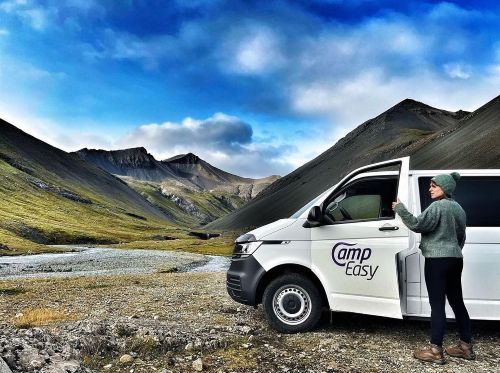 Person standing by a white CampEasy campervan parked in a scenic valley with green mountains and a winding river in the background.