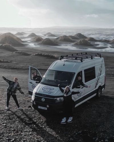 Group of three people posing joyfully next to a campervan, with dramatic sandy hills and a bright sky in the background.