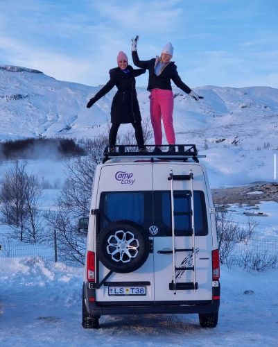 Two people standing joyfully on top of a campervan surrounded by a snowy landscape, expressing happiness with raised arms.