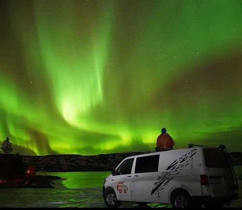 A person sitting atop a campervan watching the vibrant green aurora borealis in the night sky, with the campervan parked in a serene, rocky landscape.