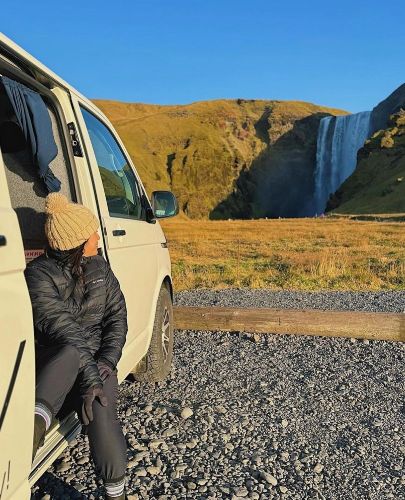 Person sitting by a campervan with a scenic waterfall in the background, enjoying a sunny day amid the vast, open landscape."
