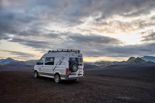 Campervan parked on a vast volcanic landscape under a dramatic sky, showcasing the rugged terrain and distant mountains.