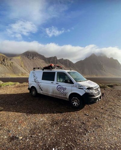 A white campervan parked on a rocky terrain with mountains and cloudy skies in the background.