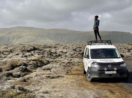 Person standing atop a campervan, surrounded by a rugged, moss-covered landscape with distant mountains under a cloudy sky.
