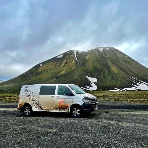 A rugged campervan stands on a road in the forefront of a mountain partially covered with snow.