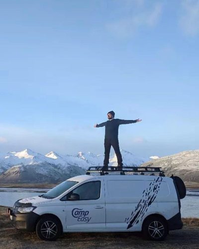 A person stands triumphantly on top of a white campervan parked against a stunning backdrop of distant mountains.