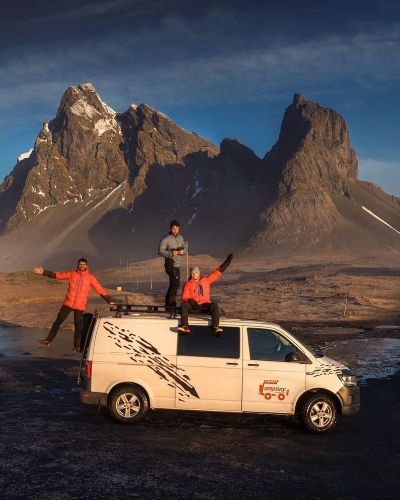 Three people are celebrating on top of a white camper van, with dramatic mountain peaks behind them.