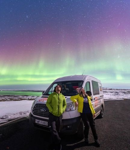 Two people are joyfully posing in front of a white camper van under a spectacular display of the Northern Lights, with a snow-covered landscape adding to the magic of the scene.