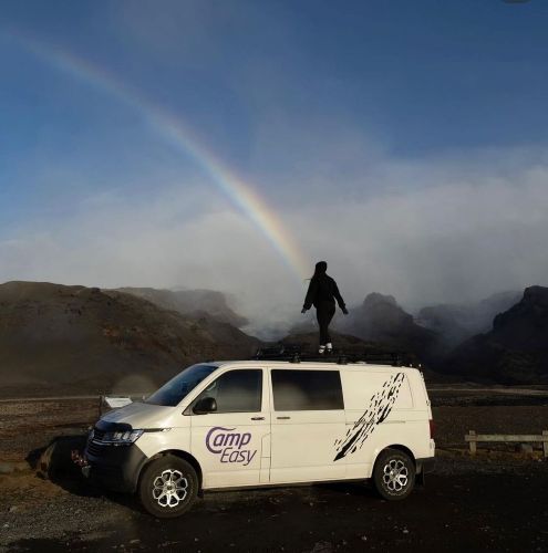 A person stands on a white camper van with a rainbow in the background. The landscape is rugged and dramatic.