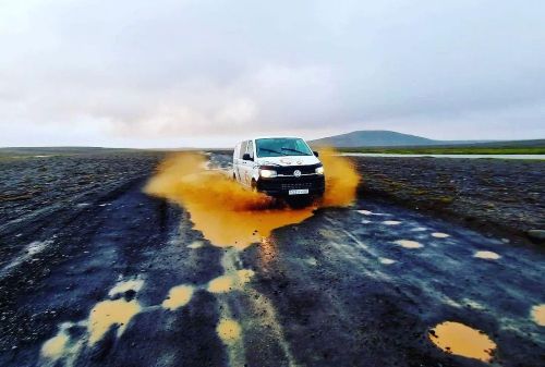 A campervan splashes through a muddy puddle on a desolate, flat landscape, sending a bright orange wave of mud into the air.