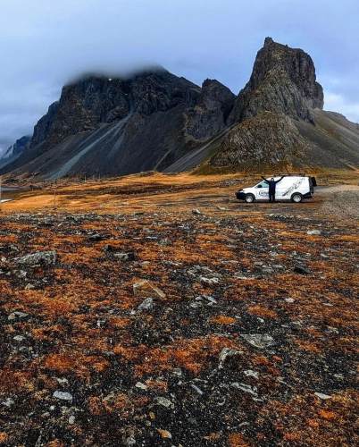 A white campervan parked in an expansive, orange-hued field with dramatic rocky mountains shrouded in clouds in the background.