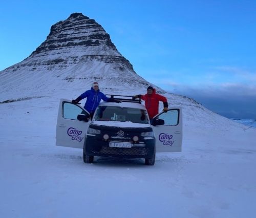 Two people are standing on the doors of a white van parked in a snowy landscape, with a distinctive, sharply pointed mountain in the background under a twilight sky.