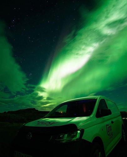 A campervan is illuminated by a vivid green aurora in the night sky, enhancing the dramatic and ethereal landscape.