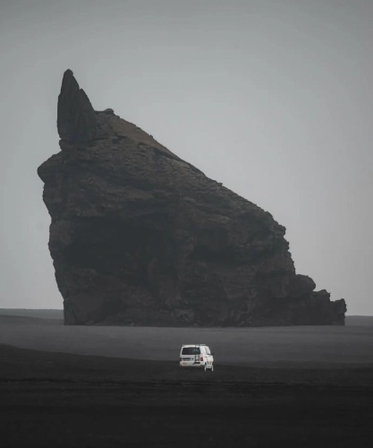 Campervan parked on a vast, dark sandy expanse, dwarfed by a massive, sharply pointed rock formation shrouded in mist.