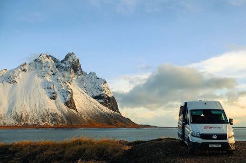 A campervan is parked by a snowy mountain and a lake. The scene is peaceful, and the light suggests sunrise or sunset.