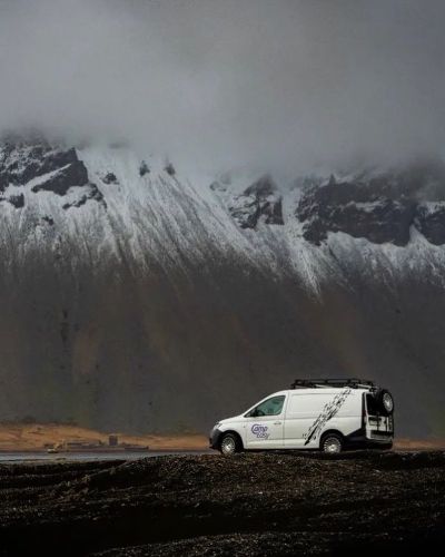 A campervan is parked on a dark gravel area, with dramatic, snow-capped mountains and moody clouds in the background.