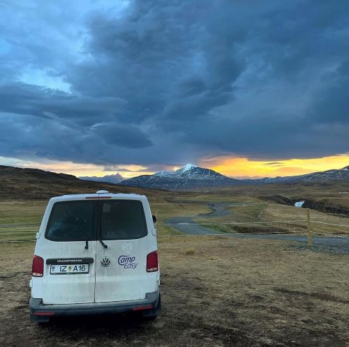 A white campervan parked on a grassy field in Iceland, facing distant mountains under a dramatic cloudy sky at sunset.