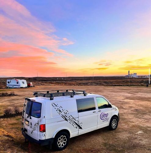A white campervan parked on an open field in Iceland, with a colorful sunset sky and another camper in the background.