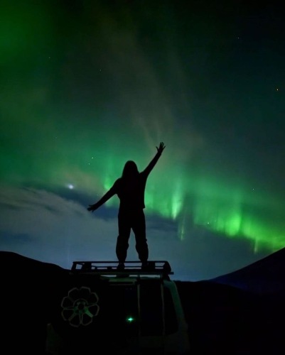 Silhouette of a person standing on top of a campervan with arms raised under the vibrant green Northern Lights in a dark night sky.