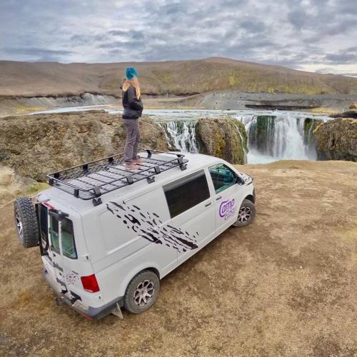 Person standing on a camper van overlooking a river and waterfall in a vast, barren landscape.