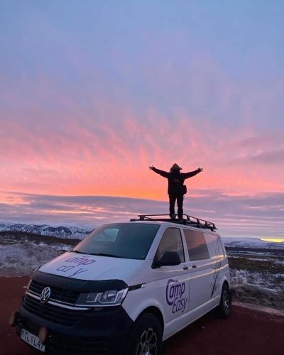 Person with arms raised standing on a camper van roof against a vivid sunset sky.