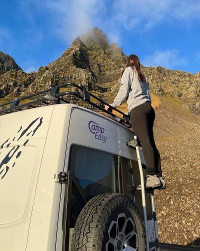 Person climbing onto the roof of a CampEasy camper van parked before a rugged mountain.