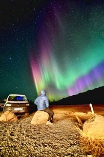 A person seated on a rock watches the colorful Northern Lights beside a parked campervan in a remote, rocky landscape at night