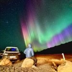 A person seated on a rock watches the colorful Northern Lights beside a parked campervan in a remote, rocky landscape at night